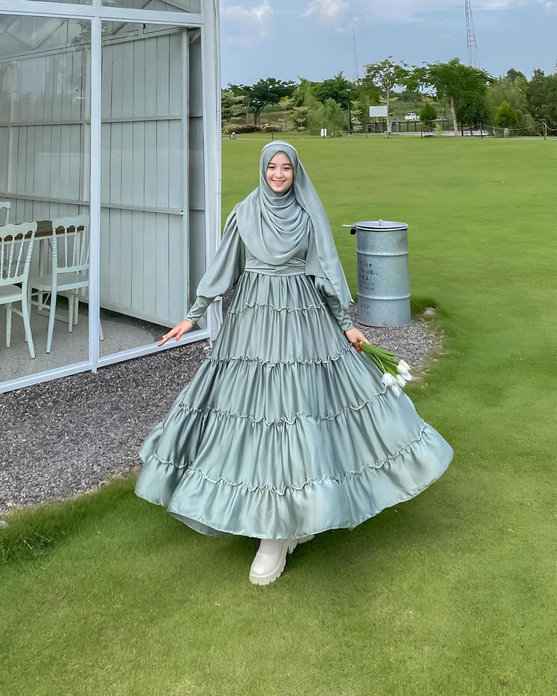 Puede incluir: Una mujer con un vestido maxi verde salvia con falda de volantes y un pa&ntilde;uelo a juego. Est&aacute; de pie en una zona de c&eacute;sped con un edificio blanco al fondo.