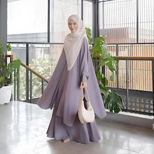May include: A woman wearing a long, flowing, light purple dress with a matching long-sleeved top. She is wearing a white headscarf and carrying a white crescent-shaped handbag.