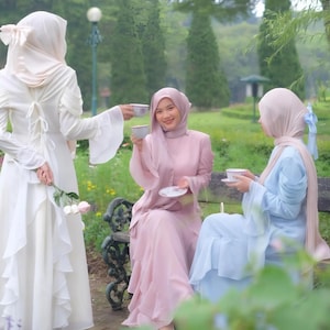 May include: Three individuals in elegant, flowing dresses and headscarves are seated outdoors, enjoying tea. The dresses are in shades of white, pink, and blue. The scene is set in a garden with lush greenery and a decorative bench.