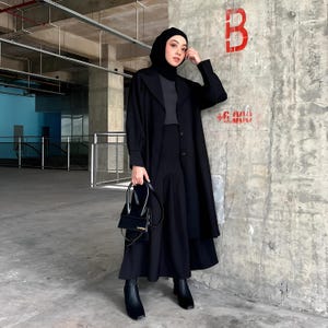 Puede incluir: Una persona con un atuendo negro, que incluye un abrigo largo, una falda y botas, está de pie en un entorno industrial. Lleva un pequeño bolso negro. El atuendo es monocromático, creando un look elegante y moderno. El fondo presenta paredes de hormigón y un acento azul.