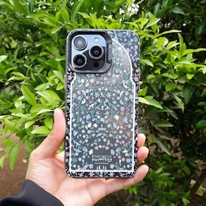 May include: A black and floral iPhone case with a clear border. The case features a pattern of small flowers in shades of blue, pink, and green. The text "SO COOL" is visible on the top right corner. The Arabic text "وردة الجنة" is at the bottom.