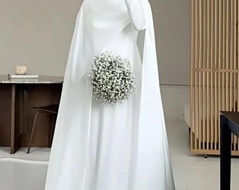May include: A white wedding dress with a long cape and a high neckline. The dress is paired with a white head covering and a bouquet of white flowers. The dress has a flowing, elegant design.
