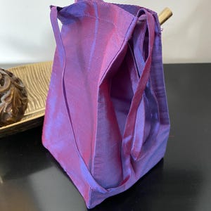 May include: A small, upright fabric bag featuring a distinct iridescent finish that shifts between shades of purple and magenta. The bag has a structured, rectangular base, a simple open top, and two thin, matching fabric handles attached to the sides.