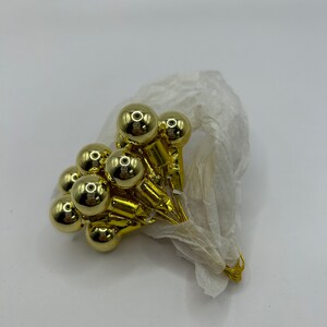 May include: A cluster of small, shiny gold-colored ornaments, each with a round shape, are arranged together. The ornaments are attached to gold-colored stems and are partially wrapped in white tissue paper. The background is plain white.