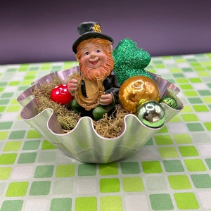 May include: A small, green leprechaun figurine holding a gold horseshoe sits in a silver, fluted metal container. The container is filled with green moss and decorated with green glitter, gold and green ornaments.