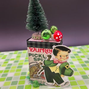 May include: A small green Christmas tree with red and green ornaments sits on top of a repurposed Durkee Pickle Spice box. A paper cutout of a leprechaun holding a hat is attached to the front of the box.