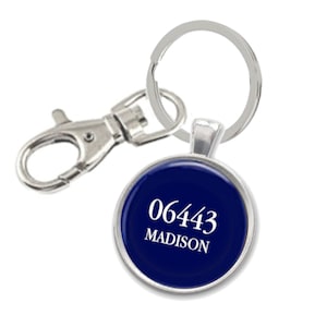 May include: A round, navy blue keychain pendant with the text "06443 MADISON" in white. The pendant is attached to a silver keyring and a silver lobster clasp.