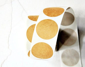 May include: A roll of circular kraft paper stickers with a peel-off backing. The stickers are a warm brown colour and are arranged in a row on a white backing. The roll is partially unrolled, revealing several stickers.
