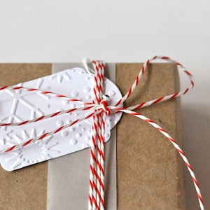 Könnte beinhalten: Ein braunes Papierpaket, umwickelt mit einer rot-weiß gedrehten Schnur, die zu einer Schleife gebunden ist. Ein weißes Geschenkanhänger mit geprägtem Schneeflocken- und Punktmuster ist mit einer kleinen silbernen Metallöse befestigt.