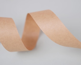 Puede incluir: Primer plano de una cinta de color beige, elegantemente curvada y ondulada. La cinta, posiblemente de papel, tiene una superficie ligeramente texturizada. El fondo es un gris suave y neutro. Ideal para manualidades.
