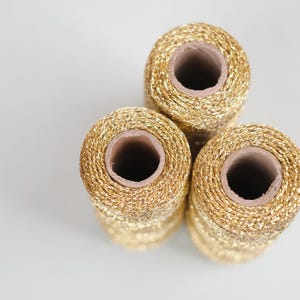 May include: Three spools of gold metallic thread are stacked on a white surface. The thread is tightly wound around a cardboard core. The spools are arranged in a triangular pattern, with the top spool slightly offset.