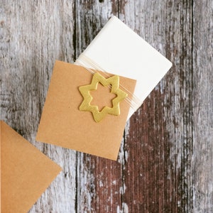 May include: A still life arrangement with a light-coloured pencil, a white square box tied with twine, and two brown square cards. A gold star-shaped ornament is placed on one of the cards. The objects are on a weathered, wooden surface.