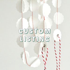 Op de afbeelding: Een decoratieve slinger bestaande uit witte, geschulpte papieren cirkels die aan elkaar zijn geregen met een dunne rode draad. De tekst CUSTOM LISTING staat in groene hoofdletters in het midden over de witte cirkels.