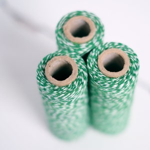 May include: Three vertical spools of baker's twine arranged in a triangular cluster. Each spool features a twisted cord pattern alternating between vibrant green and white fibres, wound around a hollow, light brown cardboard cylindrical core.