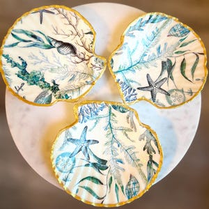 May include: Three decorative seashell-shaped dishes with a white background and a gold rim. The dishes are decorated with a blue and green watercolor pattern of seaweed, starfish, and seashells.