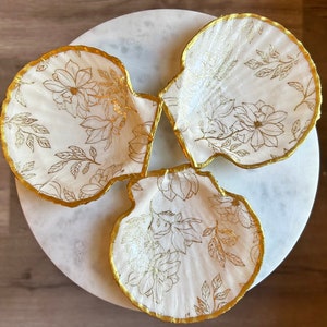 May include: Three white seashells with gold trim and a floral pattern. The shells are arranged in a circle on a white surface.
