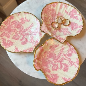 May include: Three white seashell-shaped dishes with pink floral patterns and gold trim. The dishes are arranged on a white surface. The top dish has three gold rings inside.