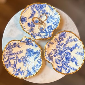 May include: Three decorative seashell-shaped trinket dishes with a blue floral pattern on a cream background. The edges are trimmed with gold. Two rings rest in one dish. Ideal for holding jewelry or small items.