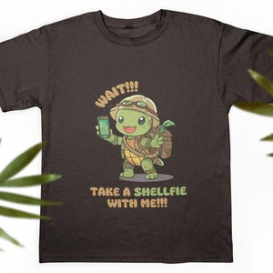 Puede incluir: Camiseta marrón oscuro con una tortuga de dibujos animados sosteniendo un teléfono. La tortuga lleva un sombrero y una mochila. El texto en la camiseta dice "WAIT!!! TAKE A SHELLFIE WITH ME!!!" Un diseño divertido y caprichoso.
