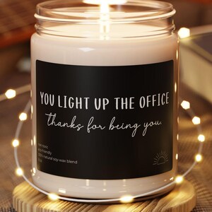 Puede incluir: Una vela en frasco de vidrio transparente con una mecha encendida, sobre una base circular de madera. La etiqueta negra presenta el texto en blanco: YOU LIGHT UP THE OFFICE, Thanks for being you, junto a texto pequeño y un icono de sol.