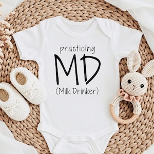 Pode incluir: Um body branco de manga curta para bebé com o texto preto "practicing MD (Milk Drinker)" na frente. O body é acompanhado por um par de sapatinhos de crochet brancos e um guizo de coelho de crochet com um anel de madeira.