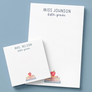 Puede incluir: Un bloc de notas rectangular y uno cuadrado, ambos blancos con texto azul oscuro. El bloc rectangular dice MISS JOHNSON fifth grade, y el cuadrado dice MRS. NELSON fifth grade. Ambos incluyen una pequeña ilustración en acuarela de libros con una manzana roja.