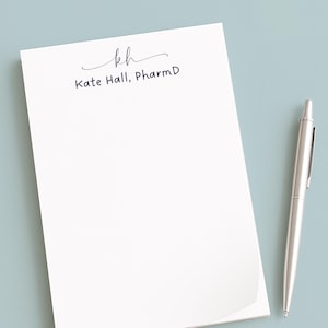 Könnte beinhalten: Ein weißer rechteckiger Notizblock mit dem schwarz gedruckten Monogramm "kh" in Schreibschrift über dem Text "Kate Hall, PharmD" in einer einfachen serifenlosen Schrift, begleitet von einem silbernen Kugelschreiber auf der rechten Seite.