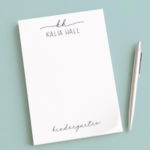 May include: A white notepad with the name "Kalia Hall" and the word "kindergarten" printed in a delicate script. A silver pen rests beside the notepad on a pale blue surface. The notepad features a personalized design.