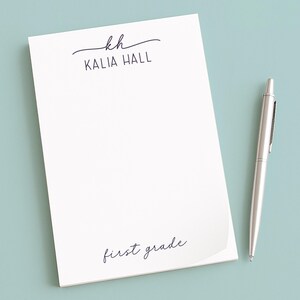 May include: A white notepad with the name "Kalia Hall" and "first grade" printed in black script. A silver pen lies beside the notepad on a light blue surface. The notepad has a simple, elegant design.