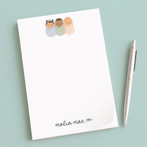 May include: A white notepad with three illustrated swaddled babies in blue, green, and peach. A silver pen rests beside the notepad on a light blue surface. The notepad has the text "malia mae, rn" at the bottom.