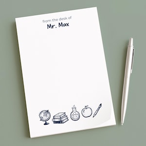 Puede incluir: Un bloc de notas blanco con el texto "from the desk of Mr. Max" e iconos escolares ilustrados. Un bolígrafo plateado descansa junto al bloc de notas sobre una superficie verde. El bloc de notas presenta un globo terráqueo, libros, un matraz, una manzana y un lápiz.