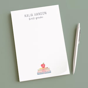 May include: A white notepad with the text "KALIA HANSON first grade" and a watercolour illustration of books and an apple. A silver pen lies beside the notepad on a green surface.