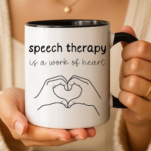 May include: A white ceramic mug with a black handle and rim. The mug features the black text "speech therapy is a work of heart" above a black line drawing of two hands forming a heart shape. The design is centred on the front of the mug.