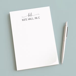May include: A white notepad with the text "kh" and "KATE HALL, PA-C" in black script. A silver pen rests beside the notepad on a light blue surface. The notepad has a curved corner.