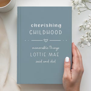 Op de afbeelding: Een stoffig blauw hardcover dagboek met witte tekst die "cherishing CHILDHOOD", een klein hartpictogram en "memorable Things LOTTIE MAE said and did" leest. Het boek ligt naast een witte koffiemok en een takje met witte gipskruidbloemen.
