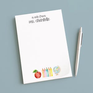 May include: A white notepad with the text "a note from ms. charlotte" in black script. The notepad features watercolor illustrations of an apple, pencils, and a globe. A silver pen rests beside the notepad on a blue surface.