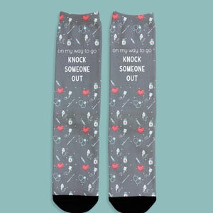 May include: A pair of gray socks with a pattern of medical symbols and red hearts. The socks have the text "on my way to go KNOCK SOMEONE OUT" in white. The socks have black toes.