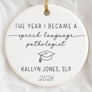 May include: A round white ceramic ornament featuring black text that reads THE YEAR I BECAME A speech language pathologist, KALLYN JONES, SLP, and 2026, with a graduation cap icon. It hangs from a gold string inside a white gift box.