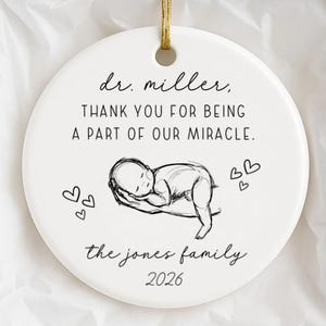 Puede incluir: Un adorno redondo de cerámica blanca con un cordón dorado, que presenta un dibujo lineal negro de un bebé sostenido en unas manos, con el texto: dr. miller, THANK YOU FOR BEING A PART OF OUR MIRACLE. the jones family 2026, y pequeños corazones.