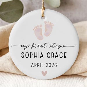 Op de afbeelding: Een ronde witte keramische ornament met een gouden ophangtouwtje. Het toont roze babyvoetjes en de tekst "my first steps", de naam "SOPHIA GRACE" en de datum "APRIL 2026", met een roze hartje eronder.