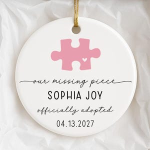 Peut inclure: Une décoration ronde en céramique blanche ornée d'un motif de pièce de puzzle rose avec un petit cœur, le texte "our missing piece", "SOPHIA JOY", "officially adopted" et la date "13.04.2027", suspendue par un cordon doré dans une boîte cadeau.