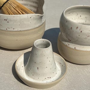 Könnte beinhalten: Ein Matcha-Set aus Keramik bestehend aus einer gesprenkelten weiß-beigen Schale mit Bambusbesen, einer passenden gesprenkelten Tasse und einem konischen Keramik-Besenhalter auf einem runden Sockel, alles mit glatter Glasur.