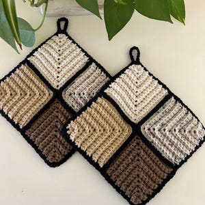 May include: Two square crochet potholders. Each potholder is divided into nine squares of varying shades of brown, beige, and cream, with a black border. A small black loop is attached to the top corner for hanging. The potholders are handmade.