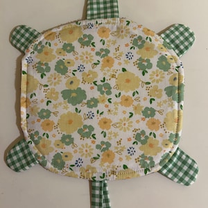 May include: A turtle-shaped fabric potholder featuring a central circular panel with a white background and a yellow and green floral pattern. The head, four legs, and tail are made from a green and white gingham checkered fabric.
