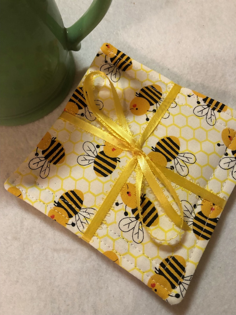 May include: A white fabric coaster with a honeycomb pattern and yellow bees. The coaster is tied with a yellow ribbon.