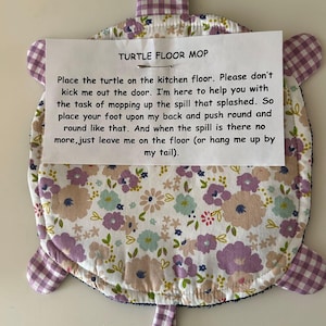 May include: A turtle-shaped floor mop featuring a white fabric top with a floral pattern in shades of purple, blue, and tan. The edges, head, limbs, and tail are made of purple and white gingham fabric. A paper note is pinned to the center.