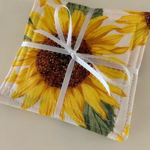 May include: A set of four square fabric coasters featuring a vibrant sunflower design with yellow petals and a brown center. The coasters are tied together with a white ribbon, creating a charming and decorative gift.