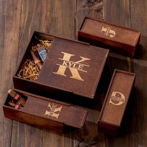 May include: A wooden gift set featuring a large box and three smaller boxes. The large box lid is engraved with the name "Kyle". The smaller boxes are engraved with the names "Johnny", "Kevin", and "Gregg". The large box contains cigars and a small bottle of liquor.
