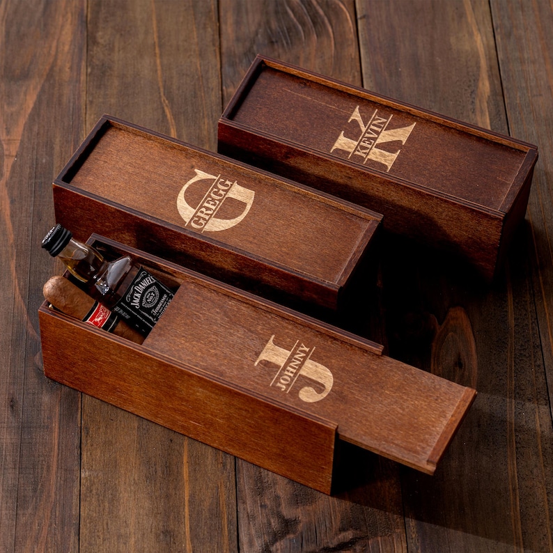 May include: Three wooden gift boxes with engraved names and initials. One box is open, revealing a miniature bottle of Jack Daniel's and a cigar. The boxes are brown and sit on a wooden surface.