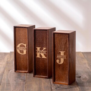 May include: Three wooden wine boxes with personalized engravings. Each box is a dark brown color and features a different name: Gregg, Kevin, and Johnny. The names are in a light brown, contrasting the darker wood.